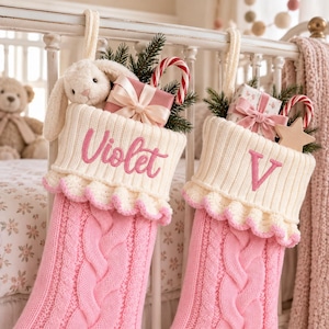 Op de afbeelding: Twee roze kerstsokken van kabelgebreid materiaal met crèmekleurige boorden en ruches. De ene sok is geborduurd met de naam Violet, de andere met de letter V. Beide zijn gevuld met knuffels, cadeautjes, zuurstokken en takjes dennennaalden.