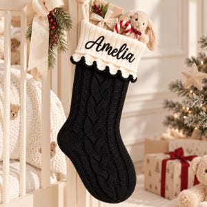 Könnte beinhalten: Ein schwarzer Weihnachtsstrumpf aus Zopfstrick mit einem cremefarbenen Umschlag mit gewelltem Rand. Der Name Amelia ist in schwarzer Schreibschrift auf den Umschlag gestickt. Ein kleiner Plüschhase und eine Zuckerstange stecken im Strumpf.