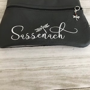 May include: A black zippered pouch with a white embroidered dragonfly and the word "Sassenach". A silver dragonfly charm hangs from the zipper pull.