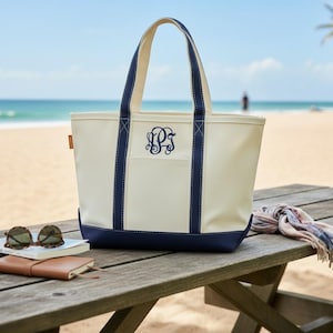 May include: A cream-colored canvas tote bag with navy blue trim and handles. The bag features a navy blue embroidered monogram on the front center. The design includes a reinforced navy blue base and two vertical navy blue straps extending from the bottom.