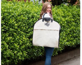 May include: A beige garment bag with black trim and black handles, featuring the embroidered monogram MGL in black thread on the front. The bag has a rounded top shape and a horizontal flap detail across the upper section of the main body.