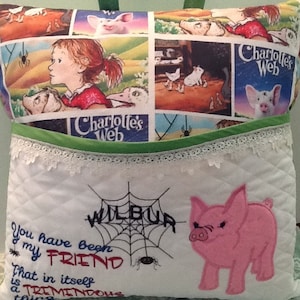 May include: A white pillow with a green pocket and a white lace trim. The front of the pillow features a pink pig, a spider web, and the text "You have been my friend. That in itself is a tremendous thing." The back of the pillow features a collage of images from the book "Charlotte's Web".