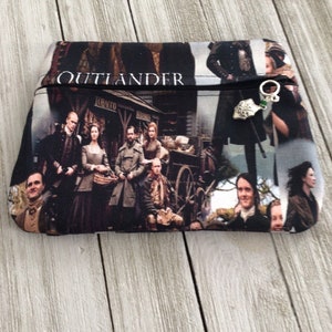 May include: A black and white zippered pouch with a collage of characters from the TV show "Outlander". The pouch has a silver zipper pull with a green charm.