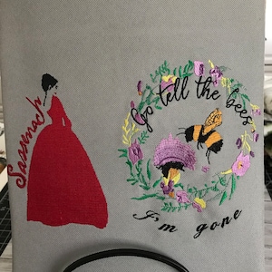 May include: A gray canvas tote bag with embroidered images. The left image is a red dress with the word "Sasenach" embroidered on the dress. The right image is a wreath of flowers with a bee and the text "Go tell the bees I'm gone".