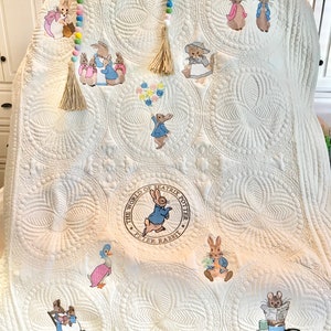 May include: White quilted baby blanket with embroidered Peter Rabbit characters and the text "The World of Beatrix Potter - Peter Rabbit".