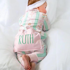 May include: A baby wearing a pink and white pajama set with a matching headband. The pajamas have a ballet-themed pattern and green stripes. The name "RUTH" is embroidered in green on the back of the pajamas.