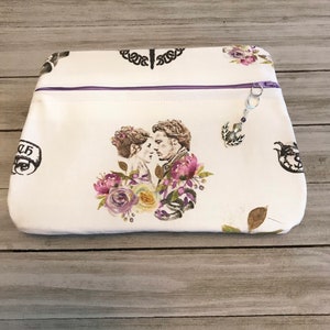 May include: White zippered pouch with a floral print and a couple's image. The pouch has a purple zipper and a silver charm with a thistle design.