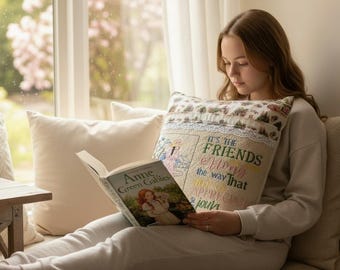 May include: A square decorative throw pillow featuring a cream-colored fabric with a lace trim and embroidered text that reads: It's the friends along the way that help us appreciate the journey. The pillow is held next to an Anne of Green Gables book.