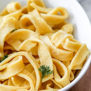 May include: A close-up shot of a white bowl filled with cooked, yellow fettuccine pasta. The pasta is arranged in loose, wavy ribbons, with a few sprigs of green parsley scattered throughout. The pasta appears to be lightly seasoned.