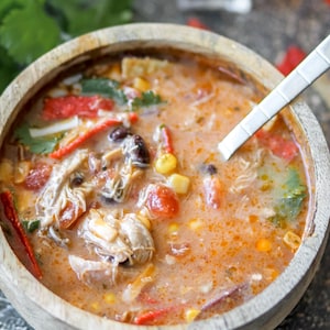 May include: A wooden bowl filled with a creamy, orange-hued soup. The soup contains chicken, black beans, corn, tomatoes, and red pepper strips. A silver spoon rests in the soup. Cilantro and red pepper flakes garnish the soup.