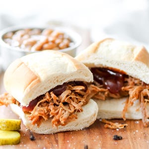 Pode incluir: Duas sanduíches de porco desfiado em pães brancos com molho barbecue, servidas numa tábua de madeira. Uma pequena tigela de feijão cozido e picles fatiados estão no fundo.