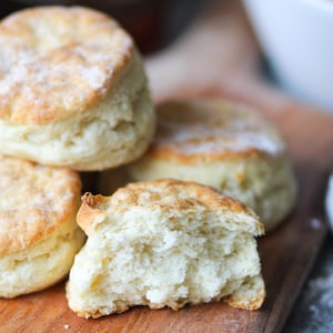 Könnte beinhalten: Nahaufnahme von mehreren goldbraunen Keksen, von denen einer aufgebrochen ist, um das flauschige Innere zu zeigen. Die Kekse liegen auf einem Holzbrett, was auf eine hausgemachte oder Bäckerei-Umgebung hindeutet. Das Bild weckt Wärme und Geborgenheit.