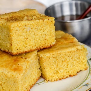 May include: Three golden-yellow cornbread squares on a floral-patterned plate. The cornbread has a slightly browned top and a textured surface. A small metal bowl and a red spoon are in the background.