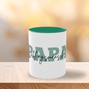 Pode incluir: Caneca de cerâmica branca com interior turquesa e a palavra "PAPA" em letras grandes turquesas. A palavra "Lieblingsmensch" está escrita em itálico abaixo. A caneca está sobre uma superfície de madeira.