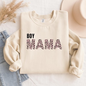 Può includere: Felpa color crema con la scritta "BOY MAMA" in nero e stampa leopardata. La felpa è piegata, con un paio di jeans blu e un cappello sullo sfondo. Il design è centrato sulla parte anteriore della maglia.