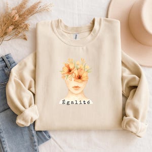 Könnte beinhalten: Cremefarbenes Sweatshirt mit einer Aquarellillustration eines Gesichts, dessen Augen von orangefarbenen Blumen bedeckt sind. Das Wort "Égalité" ist darunter in Schwarz gedruckt. Das Sweatshirt ist gefaltet, mit Jeans und einem Hut im Hintergrund.
