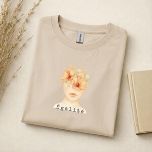 Könnte beinhalten: Beiges T-Shirt mit einer Aquarellillustration eines Frauengesichts, wobei Blumen das Haar ersetzen. Das Wort "Égalité" ist darunter aufgedruckt. Das T-Shirt ist sauber auf einer hellen Oberfläche gefaltet.