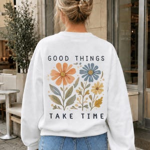 Könnte beinhalten: Ein weißes Langarm-Sweatshirt mit einer Grafik auf der Rückseite, die orange, blaue und gelbe Blumen mit grünen Stielen und Blättern zeigt. Der Text GOOD THINGS steht in Schwarz über dem Blumendesign und der Text TAKE TIME steht darunter.