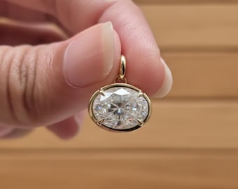 May include: A gold pendant featuring an oval-cut diamond. The diamond is set in a gold bezel and sparkles brightly. The pendant is suspended from a small gold loop. The background is a wooden surface.