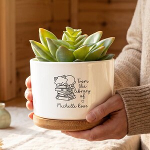 Peut inclure: Un pot de fleurs cylindrique en céramique blanche avec une base en bambou, contenant une plante succulente verte. Le pot présente un dessin au trait noir d'un chat dormant sur une pile de livres avec le texte "From the library of Michelle Rose".