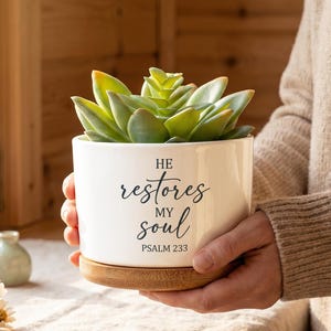 May include: A white ceramic cylindrical planter with a bamboo base, containing a green succulent plant. The front of the planter features black printed text that reads: HE restores MY soul PSALM 233, written in a mix of serif and script fonts.