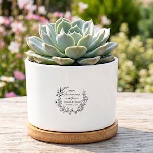 May include: A white cylindrical ceramic planter containing a green succulent plant. The planter sits on a circular bamboo base and features a black printed floral wreath design with the text Happy Anniversary, William & Olivia, October 12, 2024.