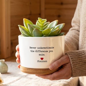 Peut inclure: Un pot de fleurs cylindrique en céramique blanche avec une soucoupe en bambou contenant une plante succulente verte. Le pot porte l'inscription noire "Never underestimate the difference you make." au-dessus d'un petit cœur rouge et du nom "Shannon".
