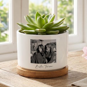Puede incluir: Una maceta cilíndrica de cerámica blanca con base de madera que contiene una suculenta verde. La maceta presenta una fotografía en blanco y negro de dos personas y el texto "Bestie Forever" impreso en una fuente cursiva en el frente.