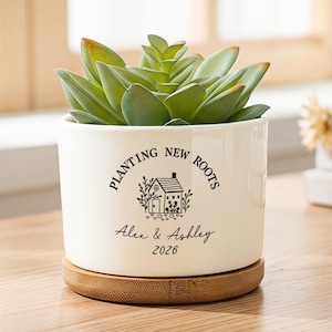 May include: A white ceramic cylindrical planter with a bamboo saucer base containing a green succulent. The planter features black text reading PLANTING NEW ROOTS, a small house illustration, and the names Alex & Ashley with the year 2026.