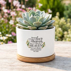 Op de afbeelding: Een witte keramische plantenbak met een vetplant, op een houten onderzetter. De plantenbak heeft de tekst "Because someone we love is in Heaven, there is a little bit of HEAVEN in our home" en een naam en data.