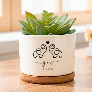 May include: A white ceramic cylindrical planter with a bamboo base containing a green succulent. The front features a black graphic of two elephants facing each other with a heart above, the text g + w, and the date 6.11.2026 printed below.