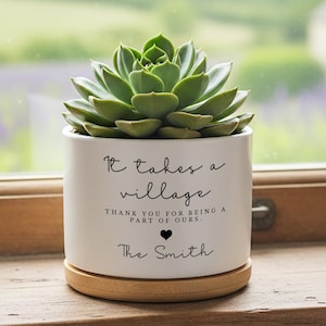 May include: A white ceramic cylindrical planter containing a green succulent. The planter features black text that reads: It takes a village, THANK YOU FOR BEING A PART OF OURS, a small black heart icon, and the name The Smith. It sits on a bamboo saucer.