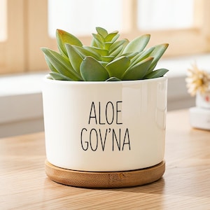 May include: A white ceramic cylindrical planter with a bamboo saucer base. The planter features the black text ALOE GOV'NA printed on the front. A green succulent plant is potted inside the container, which has a smooth, glossy finish.
