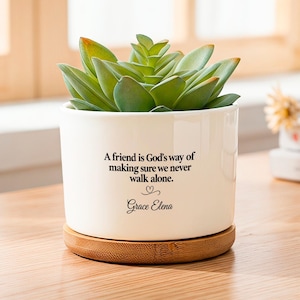 Peut inclure: Un pot de fleurs cylindrique en céramique blanche avec une base en bambou contenant une plante succulente verte. Le pot porte une inscription noire : A friend is God's way of making sure we never walk alone. Un petit cœur et le nom Grace Elena suivent.