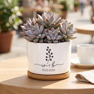 May include: A white ceramic planter containing a succulent plant, resting on a circular wooden base. The planter features a black graphic of a sprig, the letters w + b in cursive script, and the date 10.20.2024 printed on the front surface.