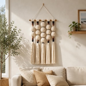 May include: A wall hanging featuring five vertical strands of cream-colored yarn spheres and tassels suspended from a horizontal wooden rod. Each strand includes dark brown and black accents between the spheres, with a long cord for wall mounting.