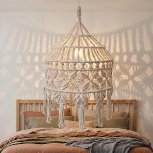 May include: Cream-colored macrame pendant light with a conical shape, hanging above a bed. The light has intricate woven patterns and decorative tassels. The bed has a wooden headboard and is covered with various colored pillows and a blanket.