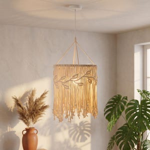 May include: A beige macrame pendant light with a leaf design. The light fixture hangs from a white ceiling fixture. The light has a cylindrical shape with a fringed bottom. The light is hanging in a room with a vase of dried plants and a large green plant.