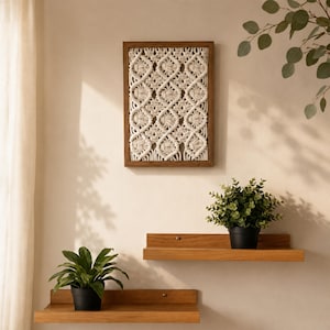 May include: A rectangular wooden frame containing an off-white macrame wall hanging with a repeating diamond knot pattern. Below the frame are two horizontal wooden floating shelves, each holding a small potted green plant in a black container.