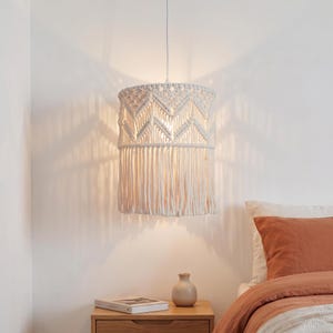 Puede incluir: Una lámpara colgante de macramé blanco. La pantalla tiene forma cilíndrica con un patrón geométrico y flecos. La luz cuelga sobre una mesita de noche con un libro y un jarrón.