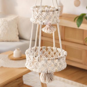 May include: A two-tiered macrame hanging basket in cream and natural tones. The baskets are round with a woven design and wooden rims. Each basket has a tassel, and the piece is suspended by thick, cream-colored cords. A decorative piece for plants or storage.