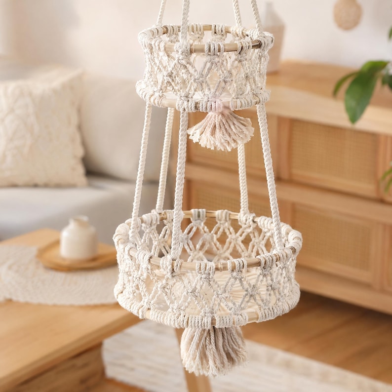 May include: A two-tiered macrame hanging basket in cream and natural tones. The baskets are round with a woven design and wooden rims. Each basket has a tassel, and the piece is suspended by thick, cream-colored cords. A decorative piece for plants or storage.