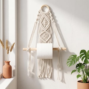 May include: A macrame toilet paper holder in natural tones. The holder features a wooden dowel for the toilet paper roll and a decorative macrame design with diamond patterns. The holder is suspended from a wooden ring.