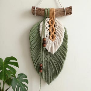 May include: Macrame wall hanging featuring a large green leaf shape with a smaller white and tan leaf accent. The design includes wooden beads and a natural wood branch for hanging. A decorative piece for home decor.