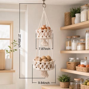 May include: A two-tiered, macrame hanging basket, 27.95 inches in height, filled with onions and potatoes. The baskets are 7.87 inches and 9.84 inches in diameter, respectively. The off-white macrame design adds a rustic touch to the kitchen.