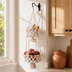 May include: A two-tiered macrame hanging basket filled with red apples and garlic and ginger. The basket is suspended from a black hook on a white wall, with a wooden cabinet and a window in the background. The basket has a natural, off-white color.