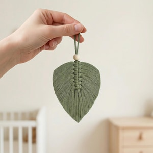 Könnte beinhalten: Handgefertigter, olivgrüner Makramee-Blatt-Dekoration mit einer Holzperle und einer Aufhängeschlaufe. Das Blatt weist detaillierte Knoten und eine strukturierte Oberfläche auf, ideal für eine natürliche Dekoration. Das Blatt ist etwa 15 cm lang.