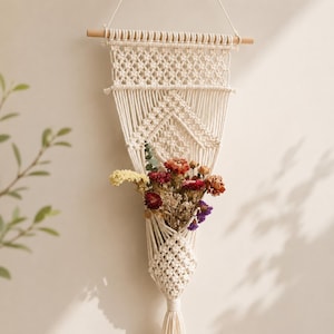 May include: Cream-colored macrame wall hanging with a wooden dowel and a woven design. The hanging features a pocket filled with dried flowers in shades of red, yellow, and purple. A decorative piece for home decor.