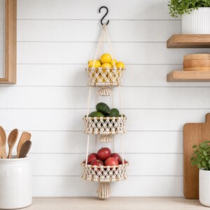 May include: A three-tiered macrame hanging basket filled with lemons, avocados, and apples. The baskets are made of woven cream-colored rope and suspended from a black hook. The fruit holder is a decorative kitchen storage solution.