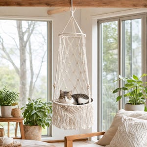 May include: A cream-coloured macrame cat bed with a woven design and fringe, suspended from a ceiling beam. A tabby cat is resting inside. The bed hangs near a window, with plants in the background, creating a cozy, natural setting.